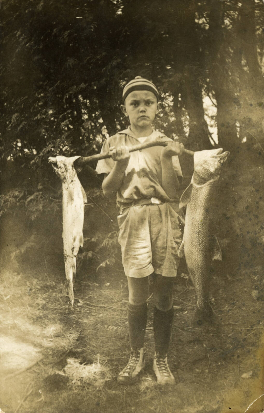 Black and white real photo postcard of a young boy holding a stick with two large fish on either end.